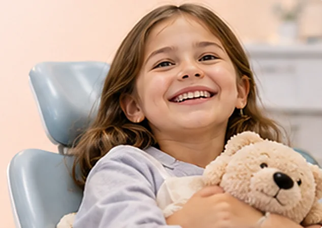 Diş koltuğunda oyuncak ayısına sarılmış gülümseyen kız çocuğu